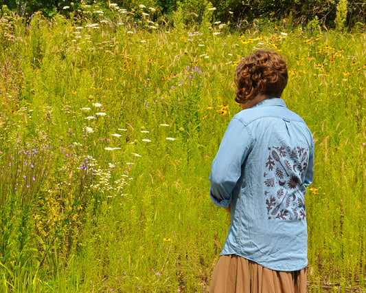 A woman stands in front of a field of wildflowers, wearing a demin shirt with a design including leaves, a camp fire, and canoe paddles.